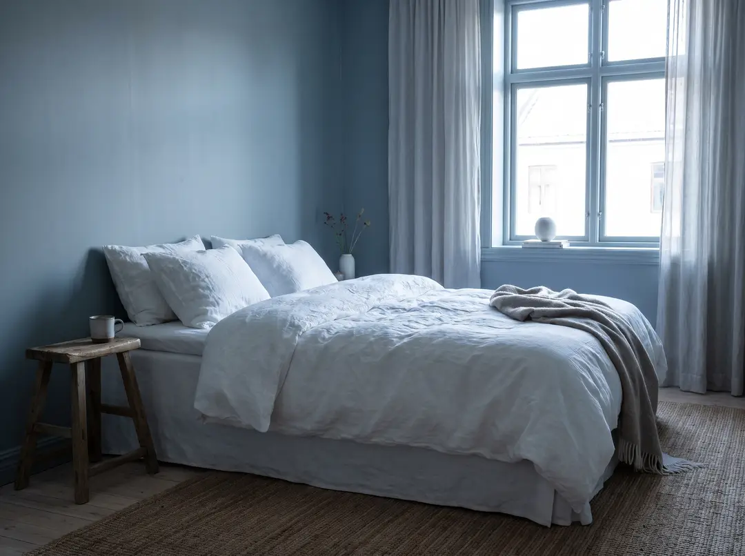 Chambre aux murs bleu gris doux, literie blanche en lin et lumière froide du nord par la fenêtre
