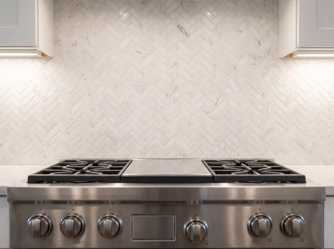 Dosseret de marbre blanc posé en chevrons derrière une cuisinière en inox, éclairage sous les armoires