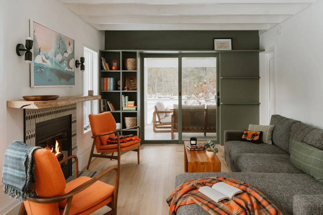 Cozy living room with lit fireplace, orange armchairs, and view of snowy forest through large glass doors