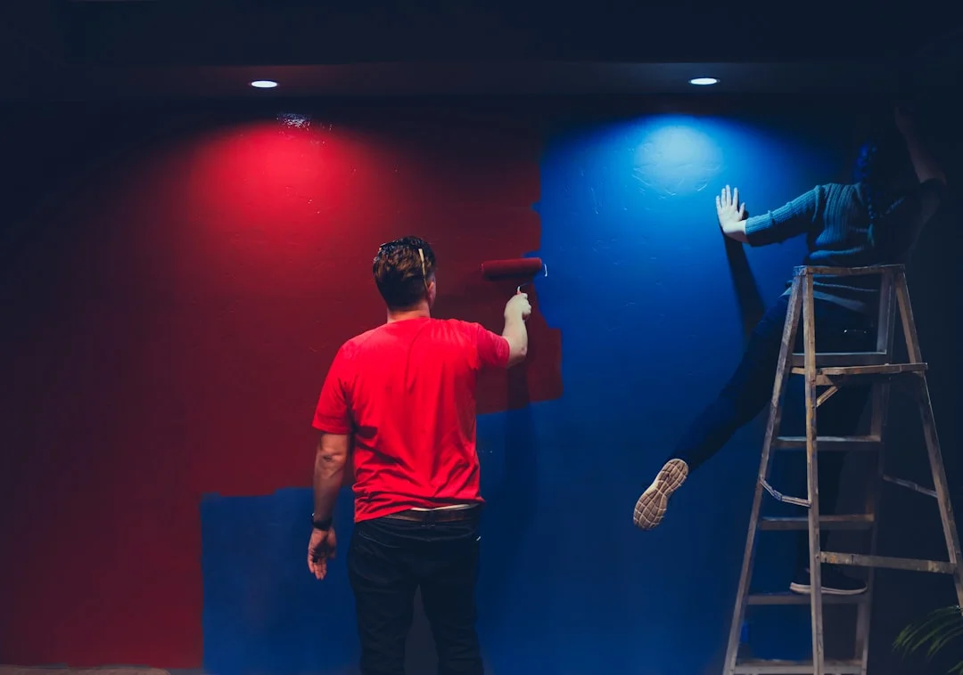 Deux personnes peignant un mur, l'un en t-shirt rouge avec un rouleau, l'autre sur une échelle, mur mi-rouge mi-bleu