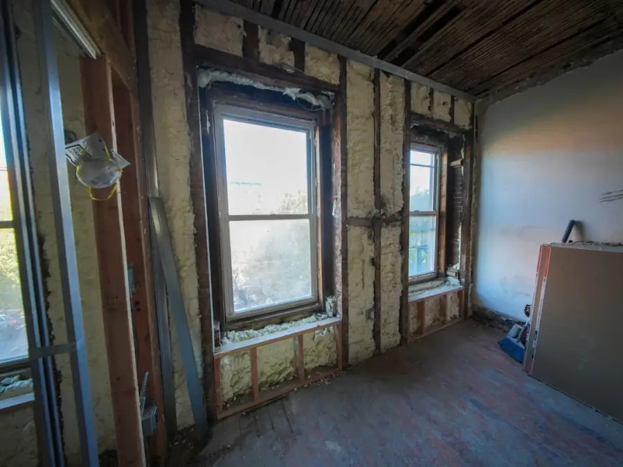 Interior room under renovation showing yellow spray foam insulation between wooden studs and two windows