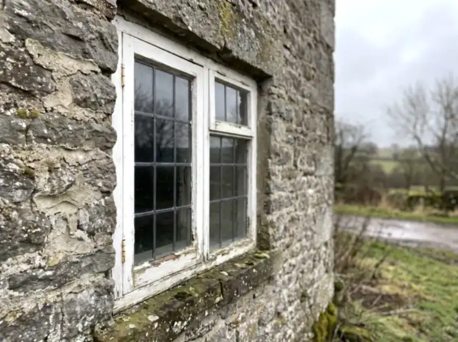 White wooden casement window in an old grey fieldstone wall, with worn mortar joints and moss on the sill.