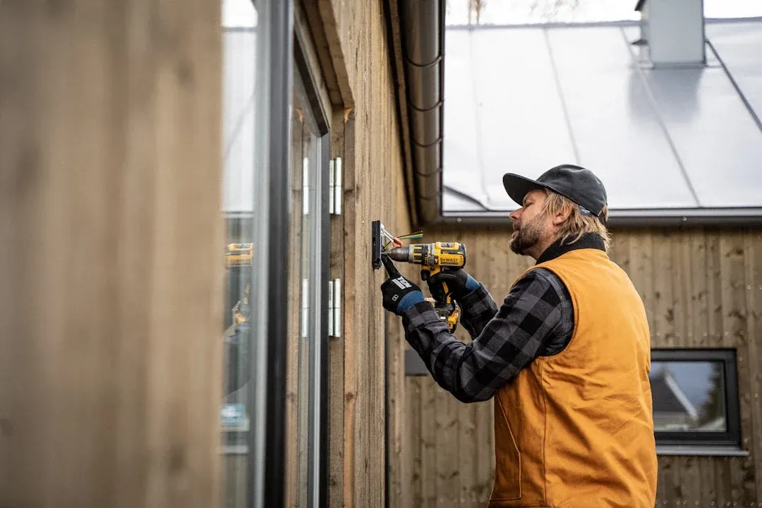 Worker in an orange vest using a DeWalt drill to install a window on a wood-frame house exterior