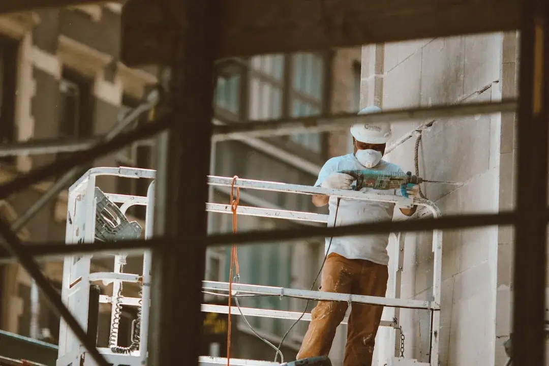 Worker in white hard hat and mask on metal scaffold using a rotary hammer drill on a stone wall