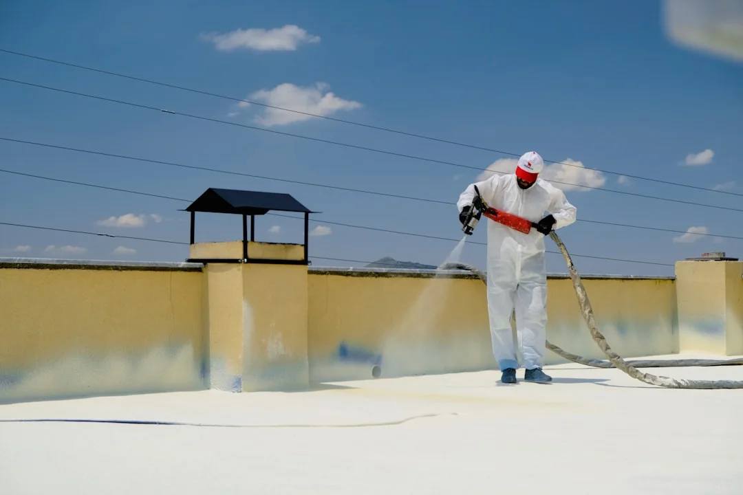 Worker in white protective suit applying insulating foam with a spray gun on a flat roof under blue sky